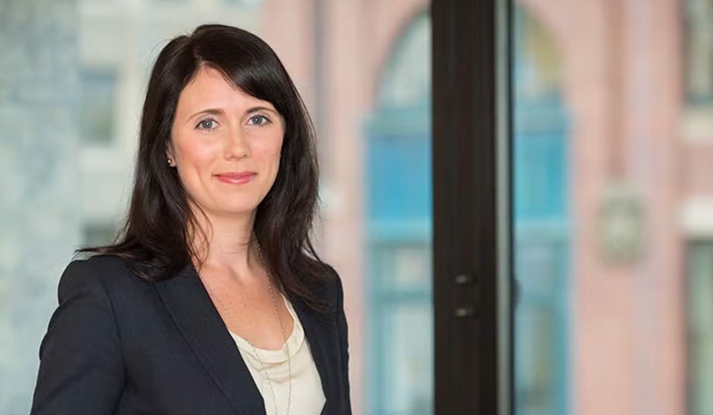 A woman in professional attire standing in front of office windows.